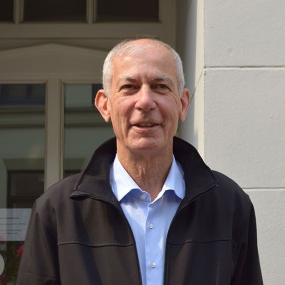Ein Mann in einem blauen Hemd und einer schwarzen Jacke steht vor einem Gebäude