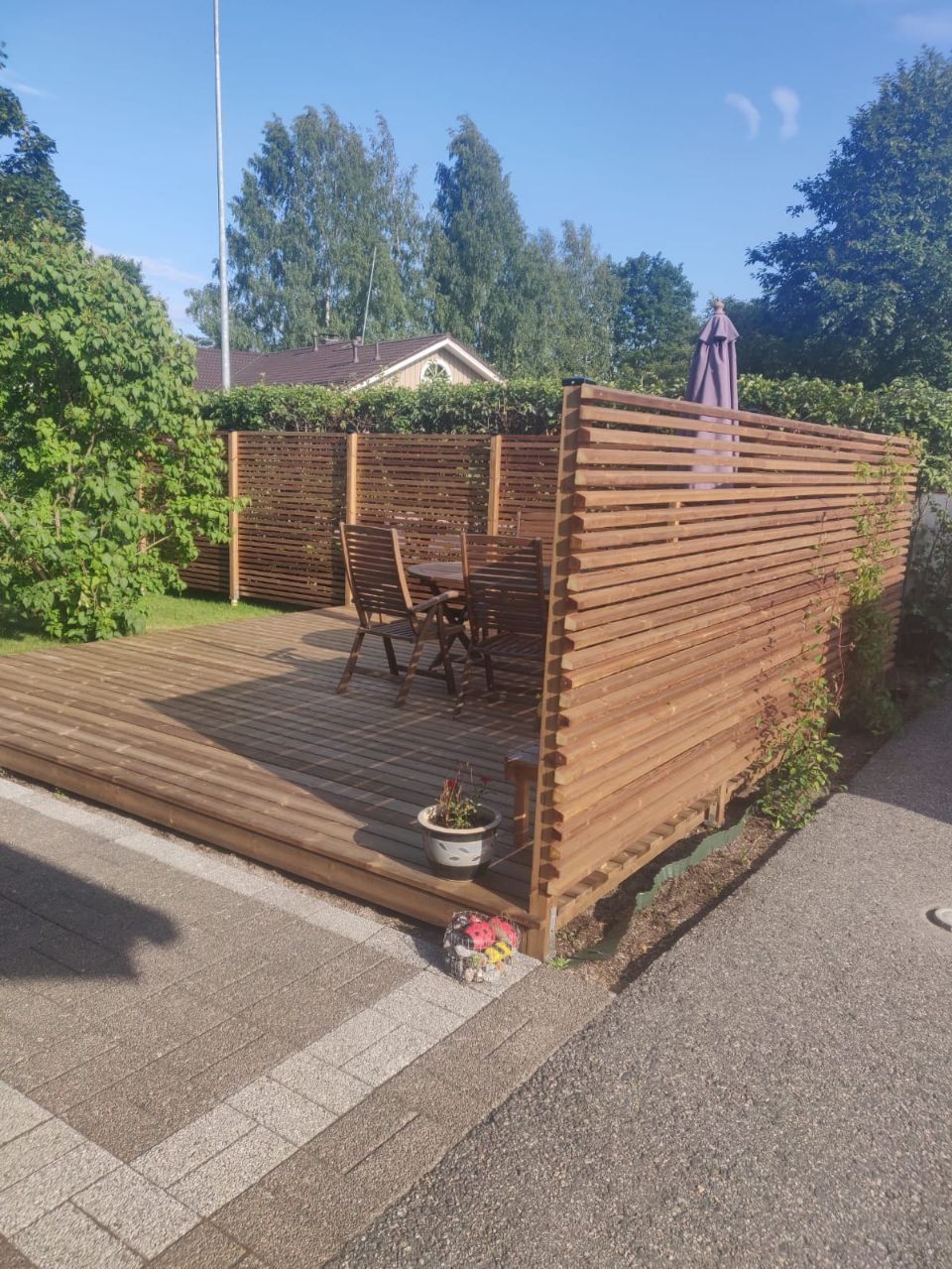 Puinen terassi, jossa on yksityisyyttä suojaava aita, pöytä ja tuolit pihalla. Aurinkoinen päivä.