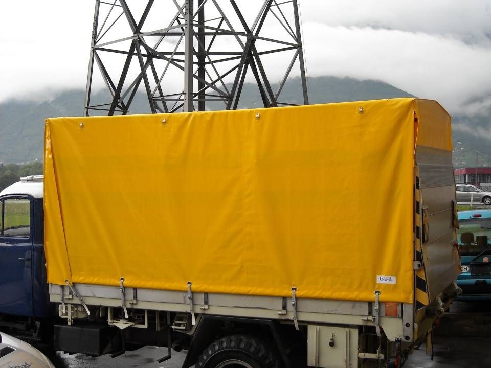 Un camion coperto da un telone giallo è parcheggiato vicino a una torre d'acciaio, con colline nebbiose sullo sfondo.