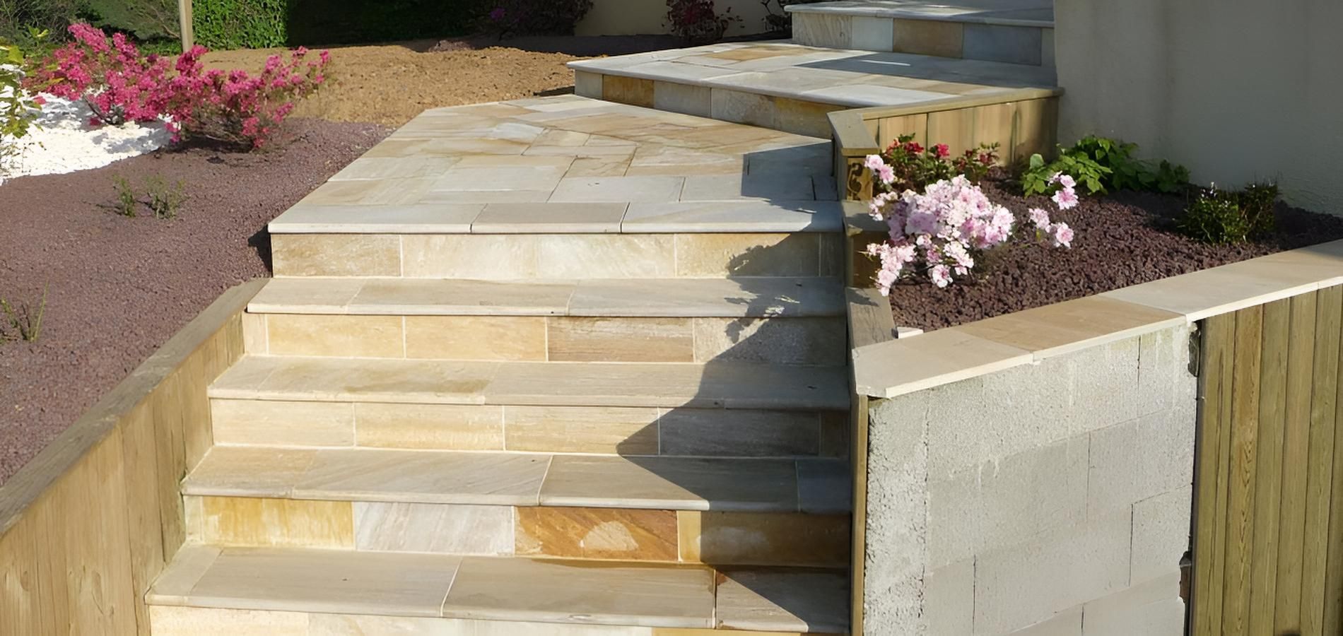 Escaliers extérieurs en grès avec murs de soutènement, bordés de parterres de fleurs roses et de terreau.
