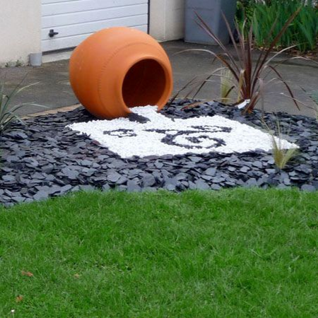 Pot en terre cuite renversé dans un jardin de rocaille, avec des pierres blanches et de l'herbe devant une maison