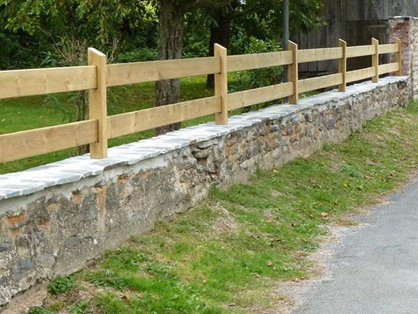 Clôture en bois sur un muret en pierre au bord d'un chemin herbeux.