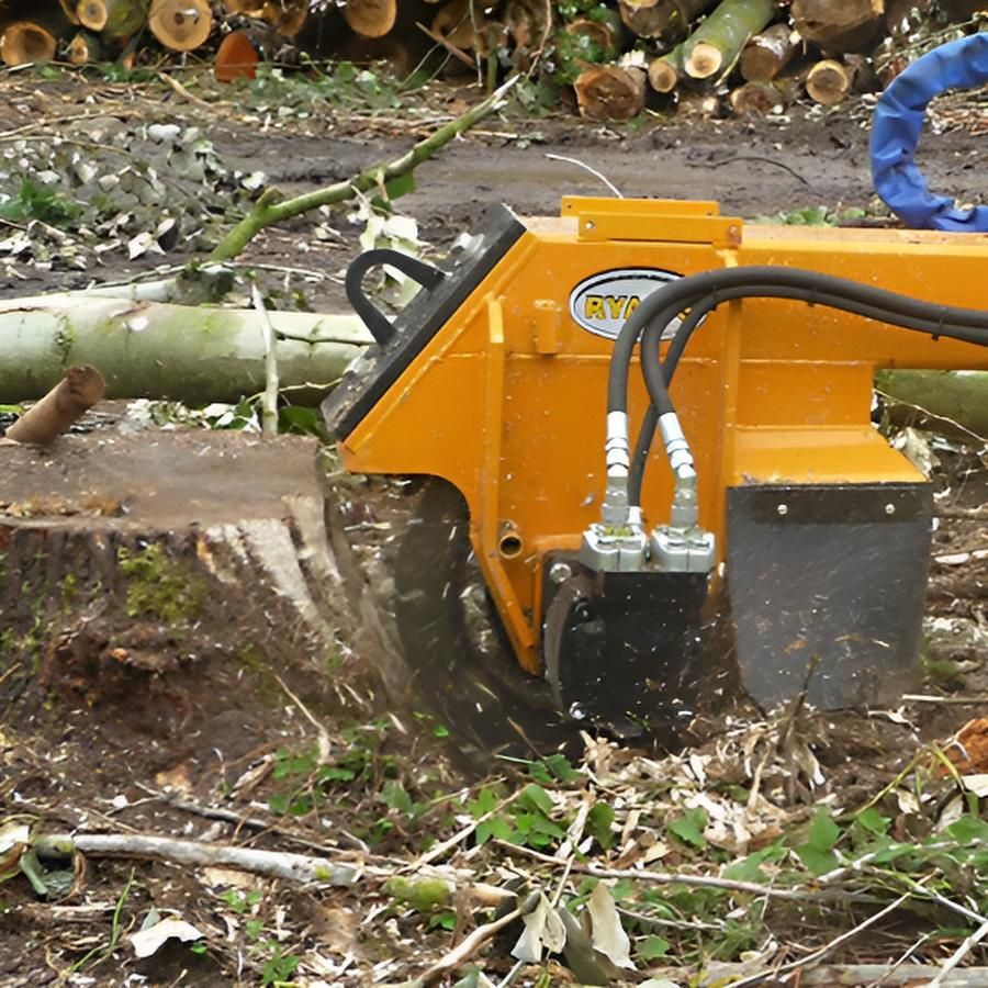 Une dessoucheuse hydraulique jaune en train d'enlever une grosse souche d'arbre dans une zone boisée avec des troncs d'arbres en arrière-plan.
