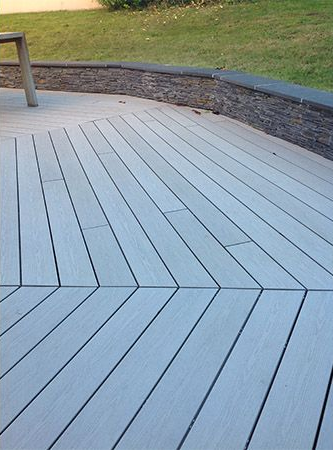Lames de terrasse composites gris clair disposées en chevrons, avec un mur de soutènement incurvé à l'aspect pierre à l'arrière.