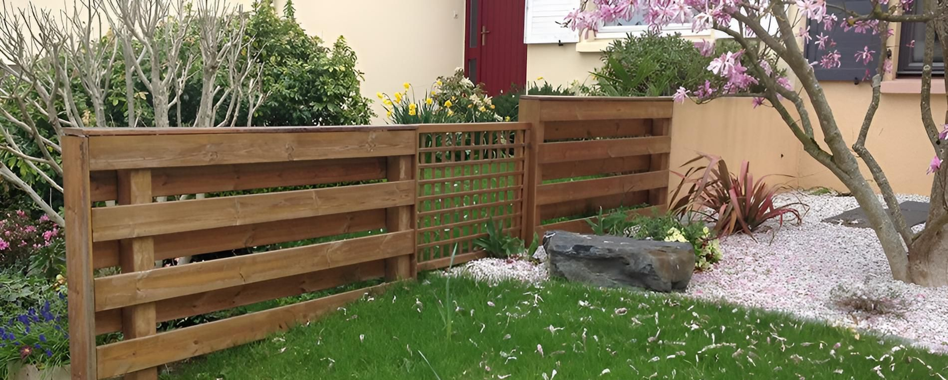 Une petite clôture de jardin en bois avec un panneau décoratif en treillis, placée dans une cour à côté d'un parterre de fleurs en gravier.
