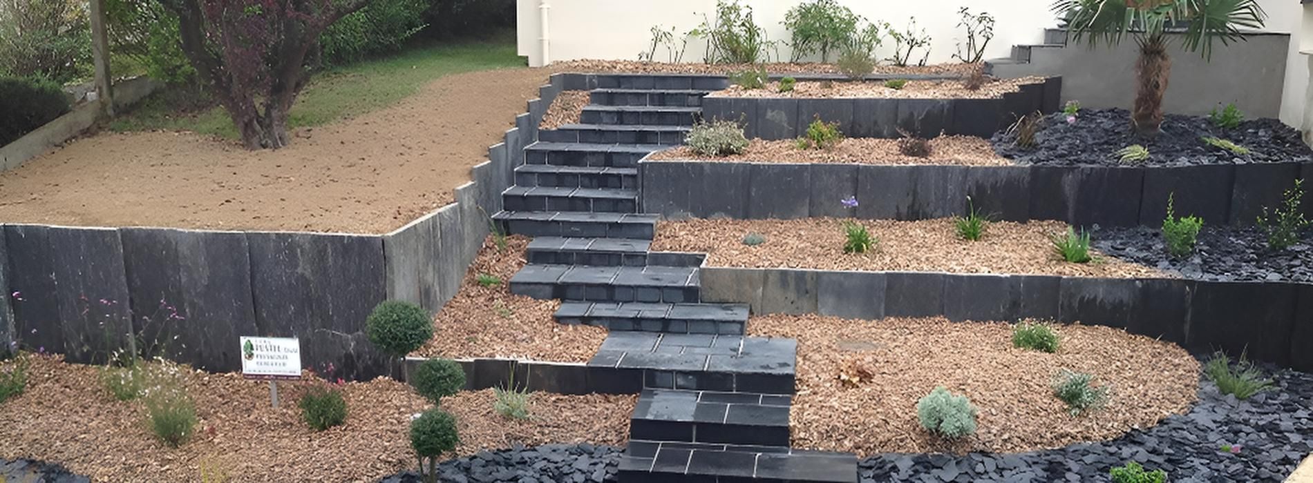 Un jardin paysager en terrasses, avec un escalier central en pierre entouré de parterres de gravier et de murs de soutènement en pierre.