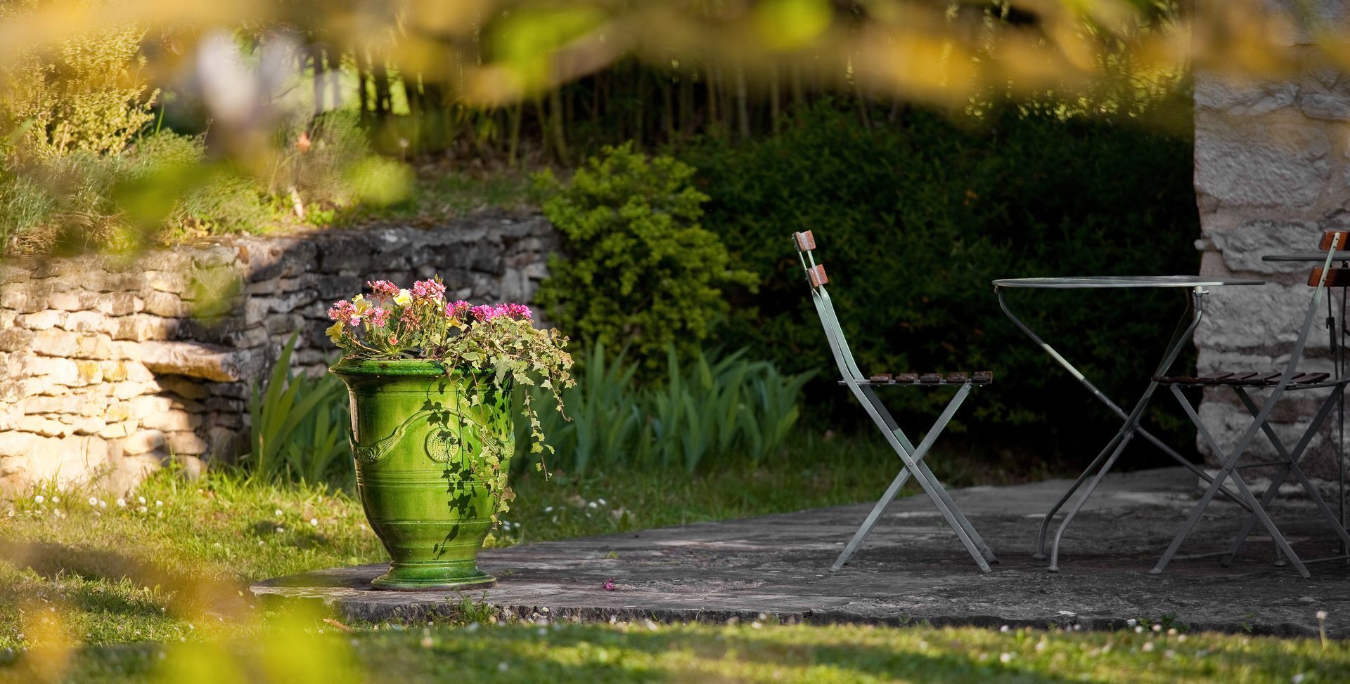 Un pot de fleurs en céramique verte est posé sur une terrasse en pierre, à côté d'une table et de chaises de bistrot, dans un jardin luxuriant.