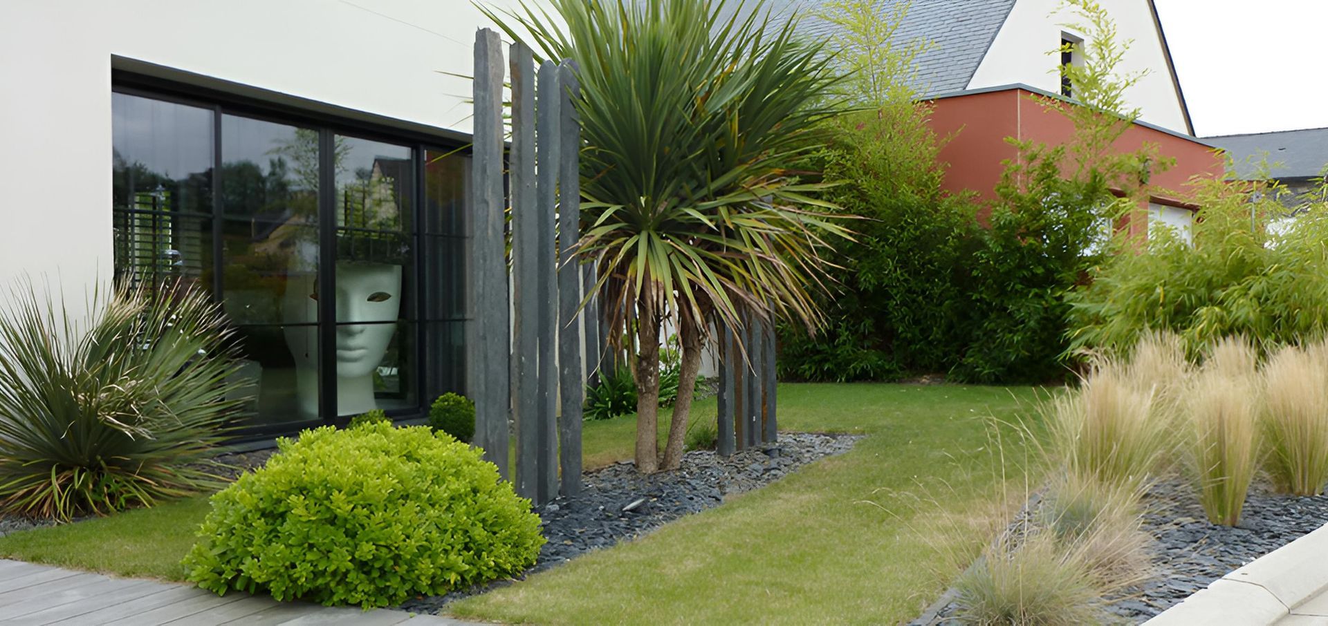 Extérieur de maison moderne avec pelouse verte, graminées ornementales, une plante ressemblant à un palmier et des piliers verticaux en pierre gris foncé.