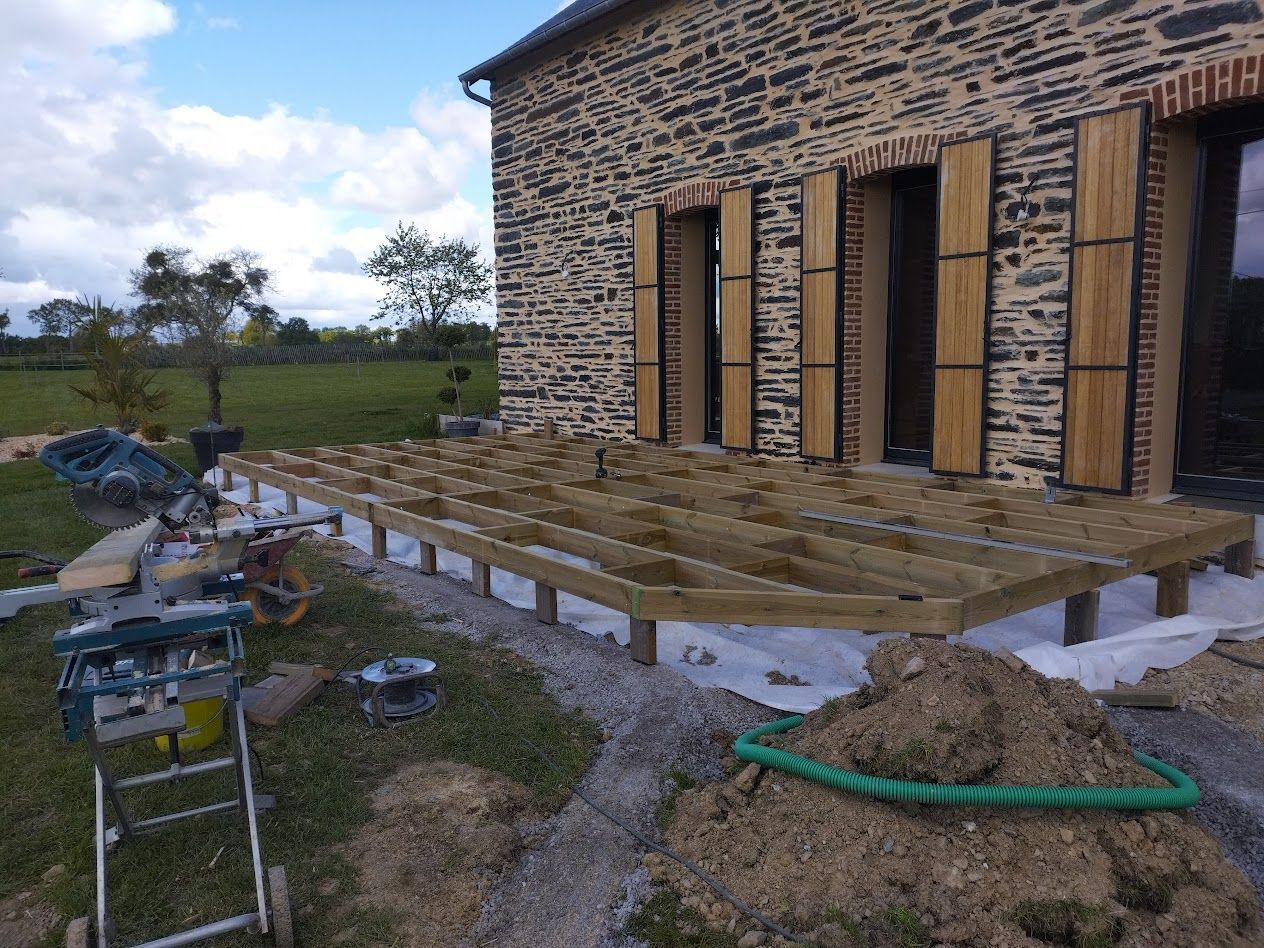 Terrasse en bois en construction à côté d'une maison en pierre, avec outils et matériaux sur un terrain herbeux.