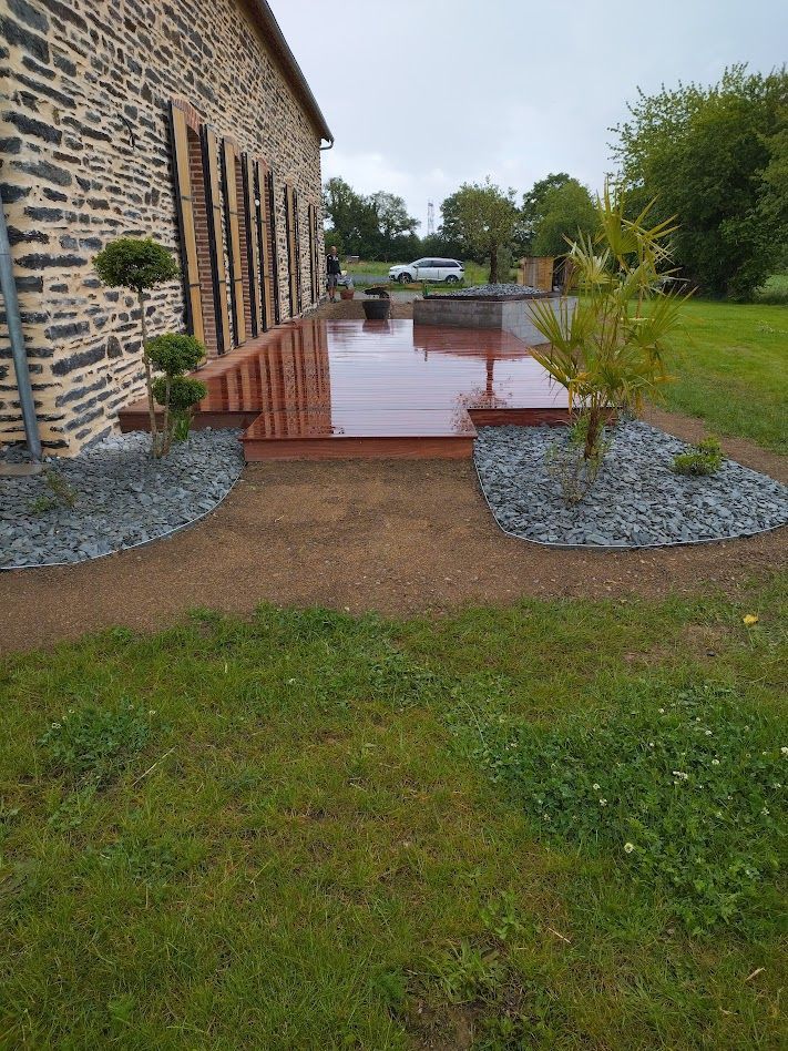 Bâtiment en pierre avec terrasse rouge donnant sur un chemin de gravier mouillé et une cour gazonnée après la pluie