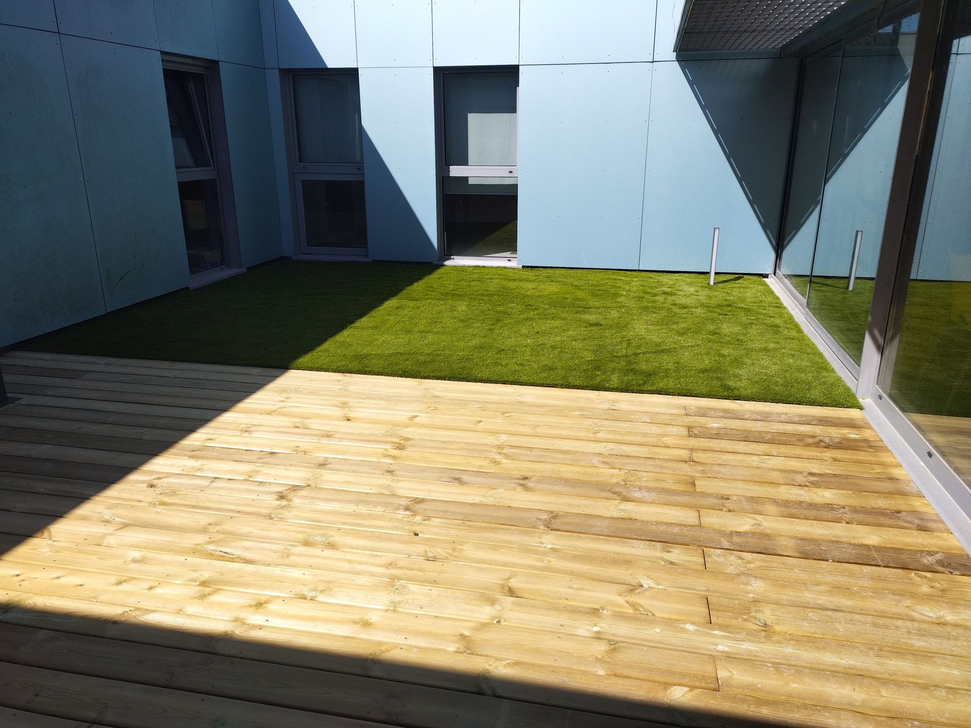 Petite cour avec terrasse en bois, pelouse verte et murs bleu clair baignés de soleil