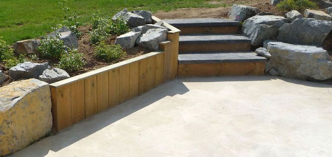 Terrasse en béton avec murs de soutènement en bois, aménagement paysager en pierre et marches menant à une pelouse.