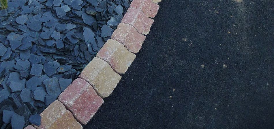 Une bordure incurvée composée de pavés rouges et beiges sépare un lit de roches d'ardoise sombre d'un chemin en asphalte noir.