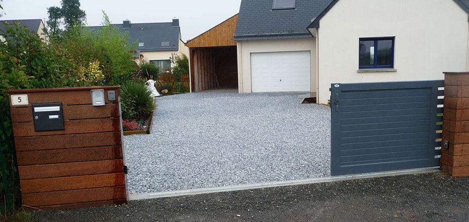 Une allée grise menant à une maison moderne, un abri voiture et un portail en bois, dans un cadre résidentiel de banlieue.