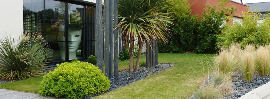 Un jardin moderne avec des graminées ornementales, un palmier, un petit arbuste vert et une grande sculpture de masque blanc dans une fenêtre.