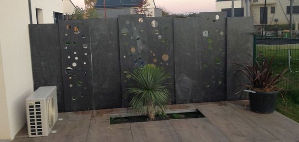 Un paravent sombre à panneaux verticaux, orné de découpes circulaires éparses, donne sur une terrasse avec un petit palmier et une plante en pot.