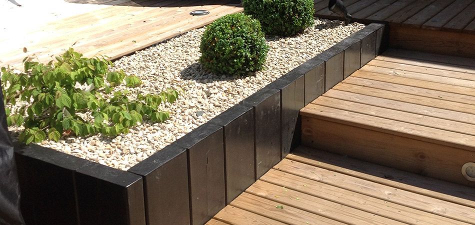 Un parterre surélevé recouvert de gravier blanc, agrémenté de deux petits arbustes ronds et de plantes vertes feuillues, jouxte une terrasse en bois et un escalier.