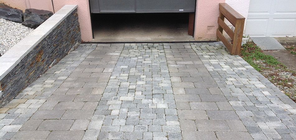 Une allée en pierre grise avec un motif central de blocs texturés, menant à une porte de garage, flanquée d'un muret et d'une clôture en pierre.