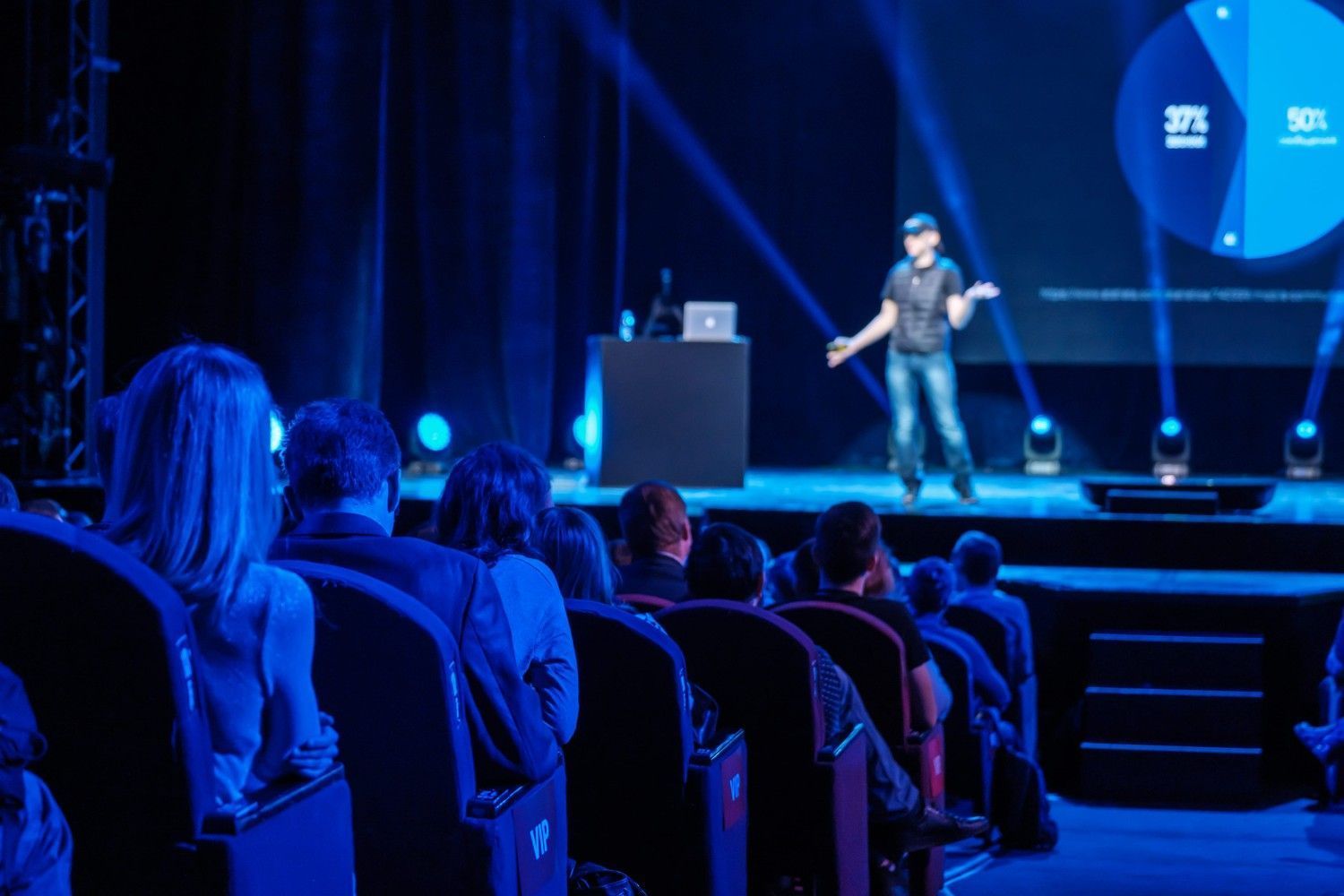 Un orador realiza una presentación en el escenario ante un público en un auditorio con iluminación tenue y tonos azules.
