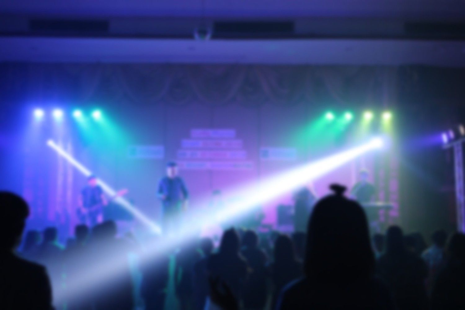 Un concierto en un escenario con brillantes haces de luz azul y verde que iluminan la silueta del público.
