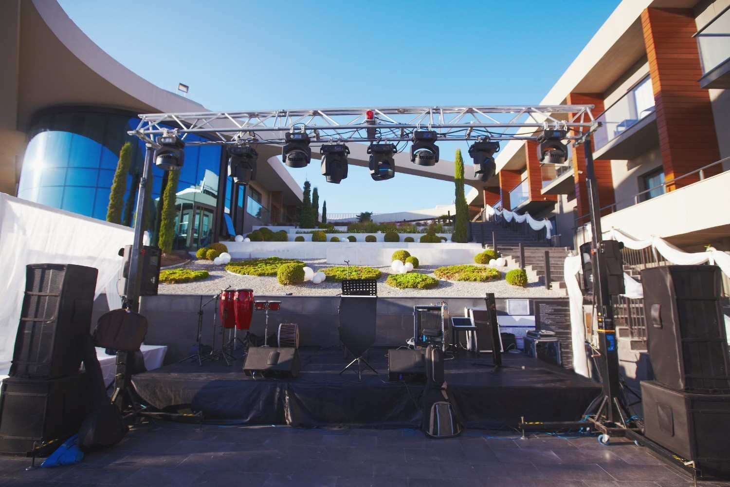 Un escenario al aire libre con altavoces, una batería y una estructura metálica con iluminación cenital.