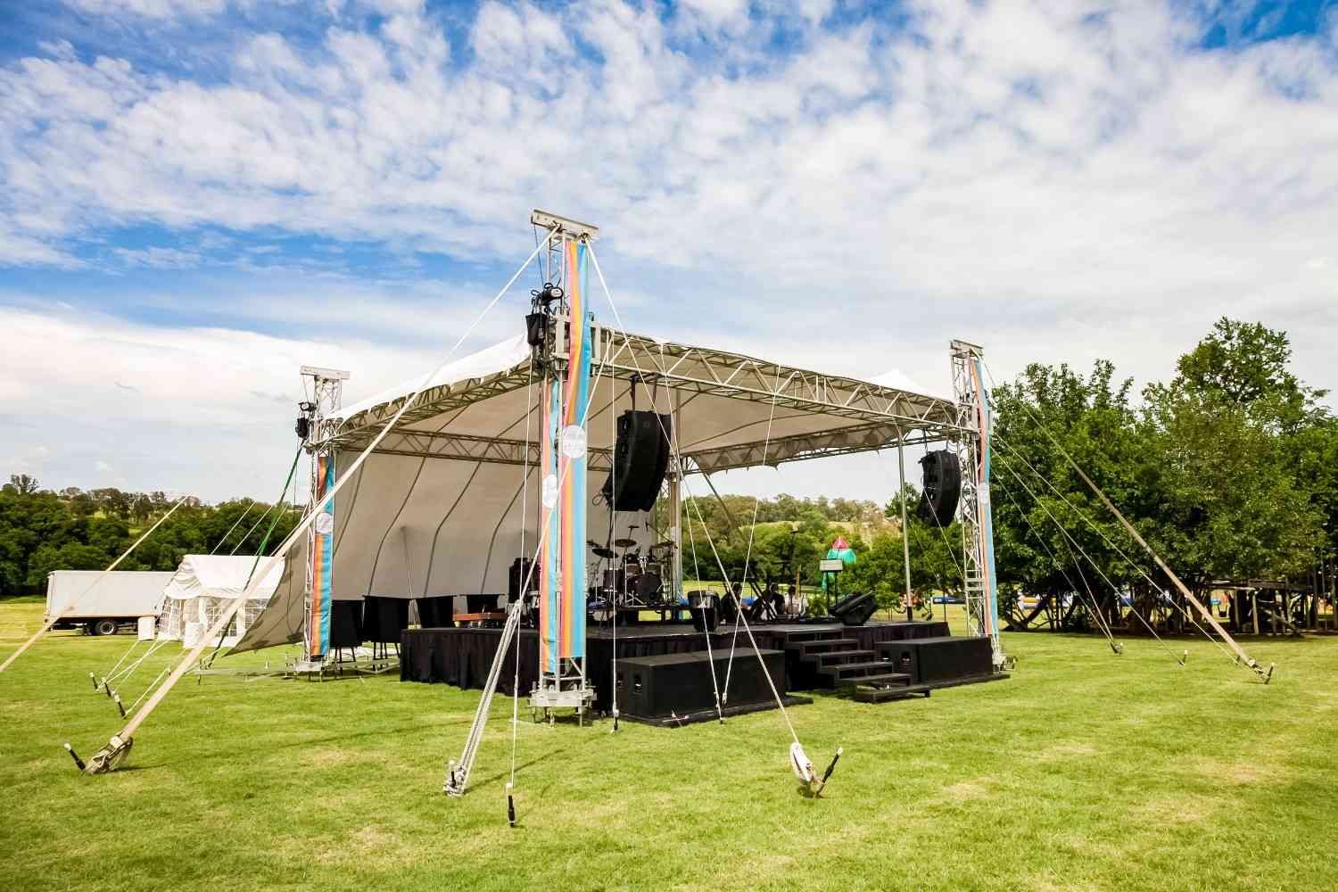 Un escenario portátil al aire libre con equipo de iluminación y sonido instalado en un campo de hierba bajo un cielo nublado.