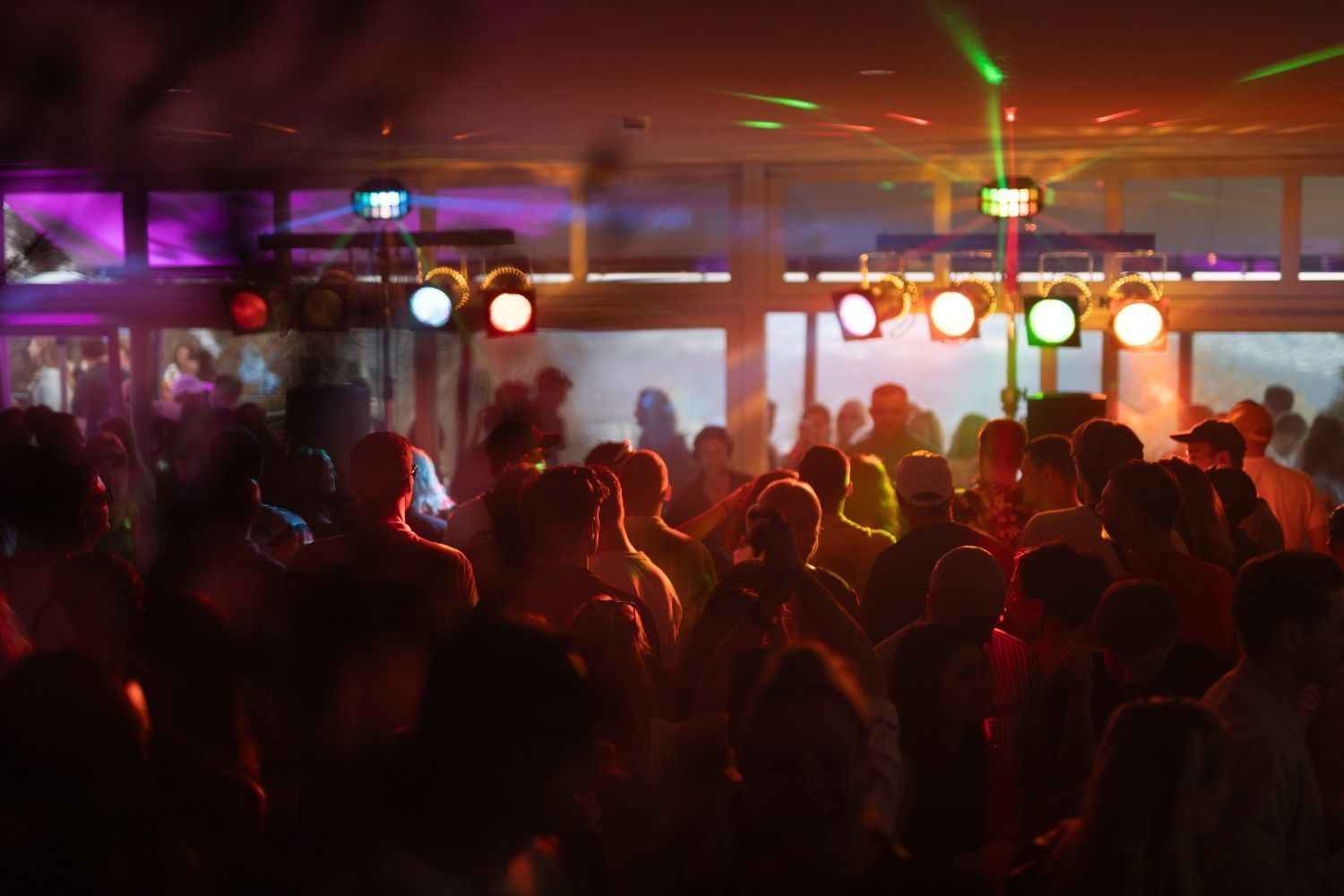 Una multitud de personas bailando en una fiesta en un espacio cerrado con luces de escenario brillantes y focos de colores.