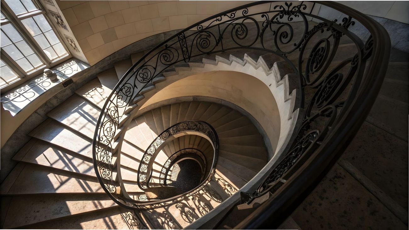 Escalier en colimaçon avec rampe en fer ornée, lumière naturelle projetant des ombres.