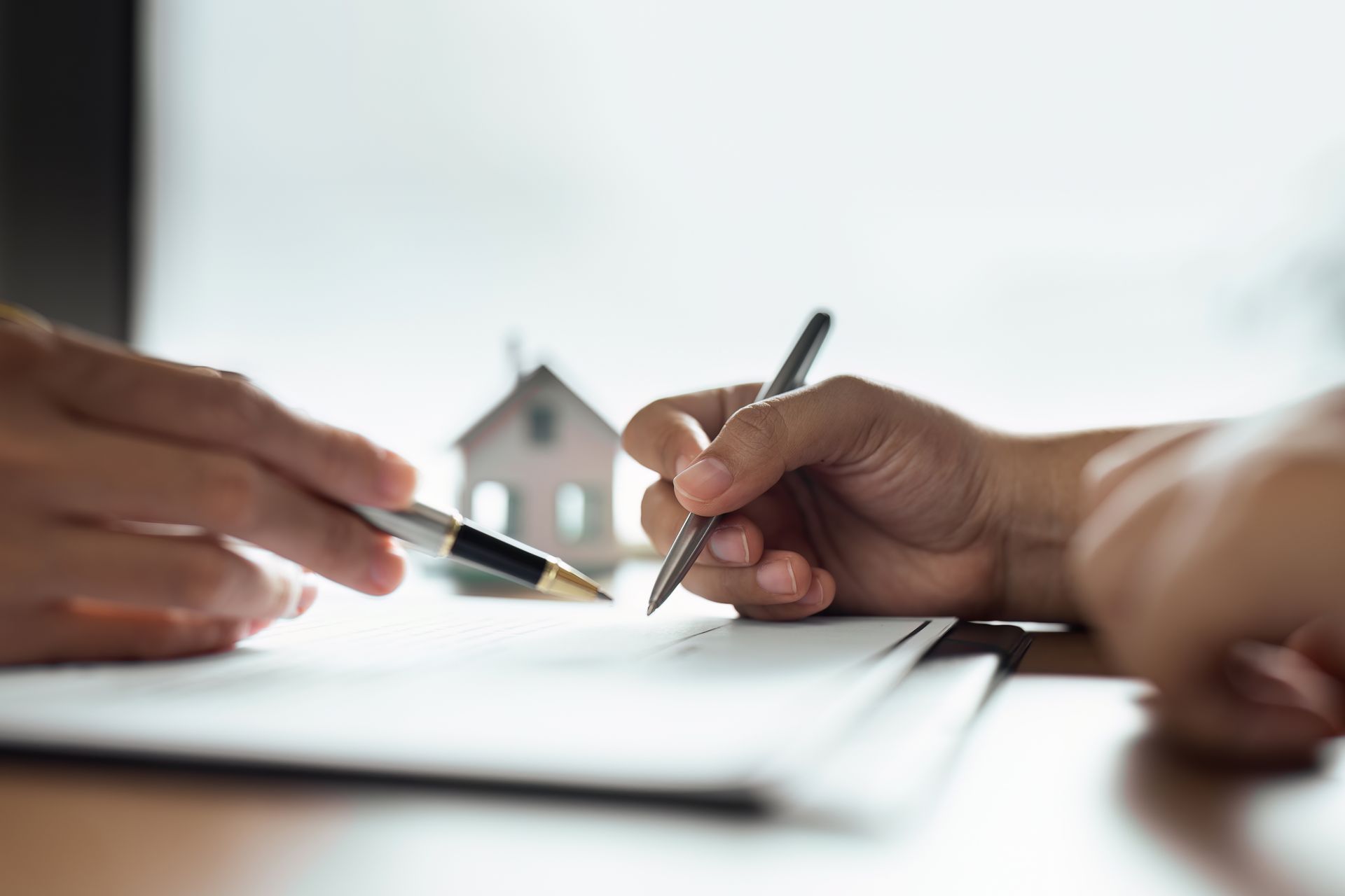 Deux mains face à face tenant chacune un stylo avec silhouette de maison en arrière-plan