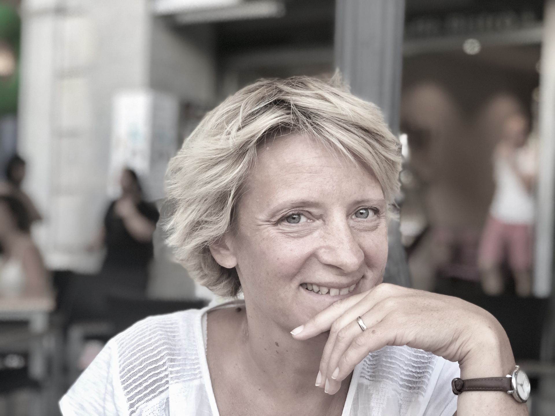 Une personne aux cheveux blonds courts porte une chemise blanche et sourit en posant son menton sur sa main à la terrasse d'un café.