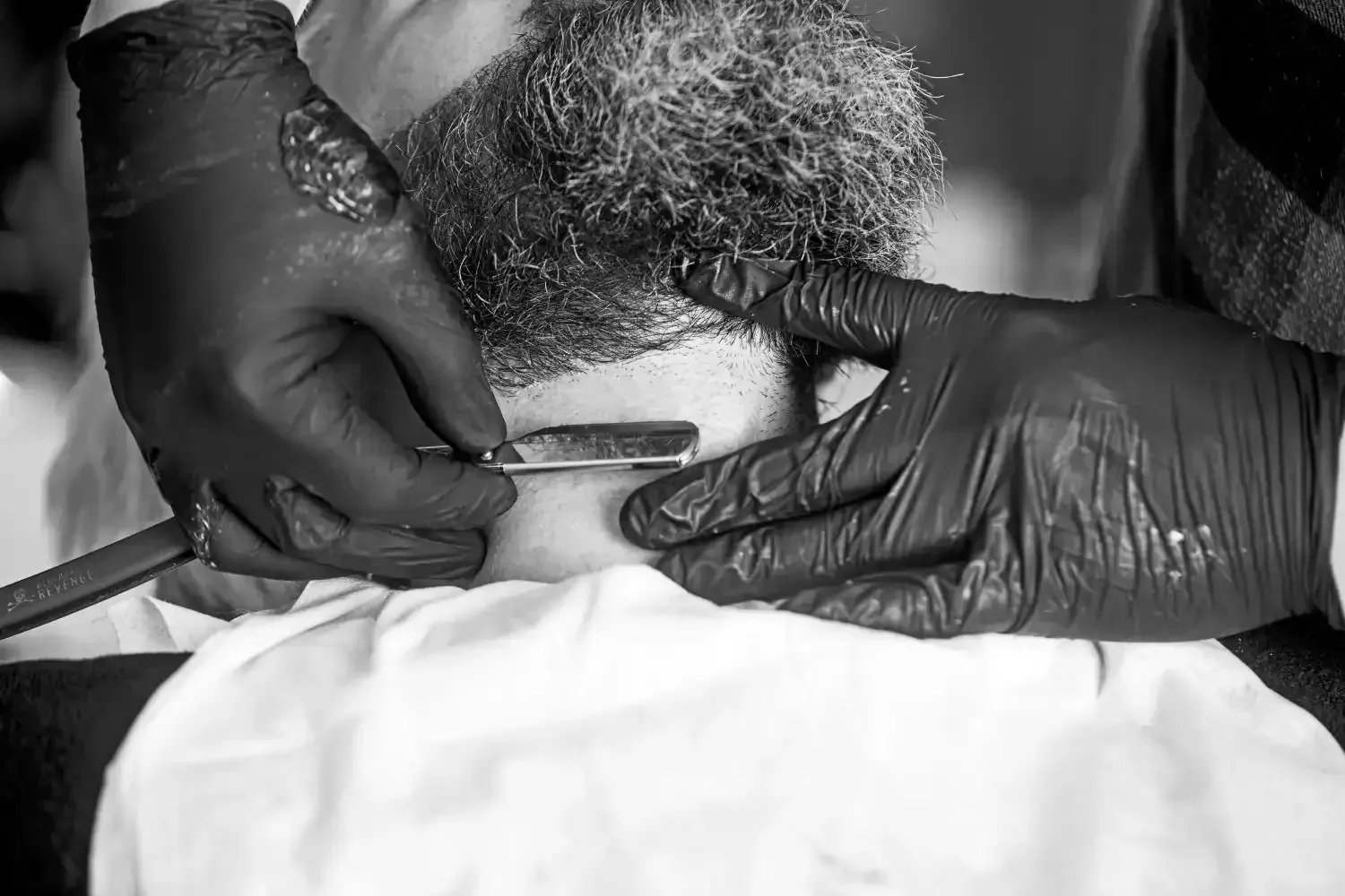 El barbero afeita la barba de un cliente con una navaja de afeitar; manos enguantadas con guantes negros y una toalla blanca.