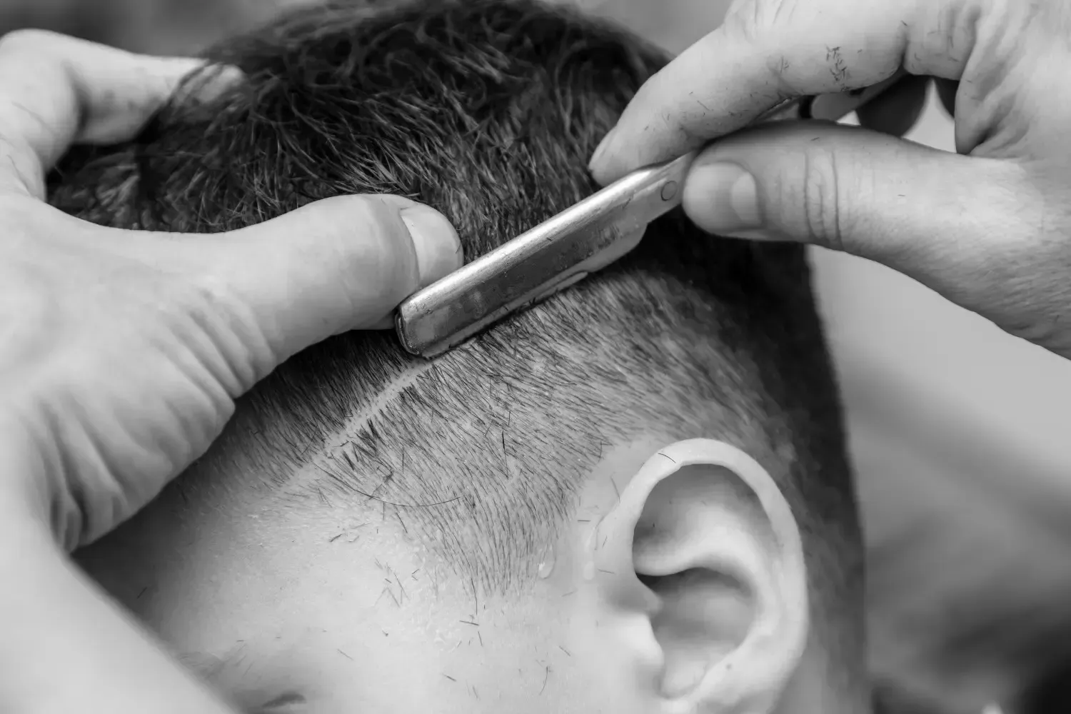 Un barbero afeita la línea del cabello de un cliente con una navaja de afeitar; primer plano en blanco y negro.