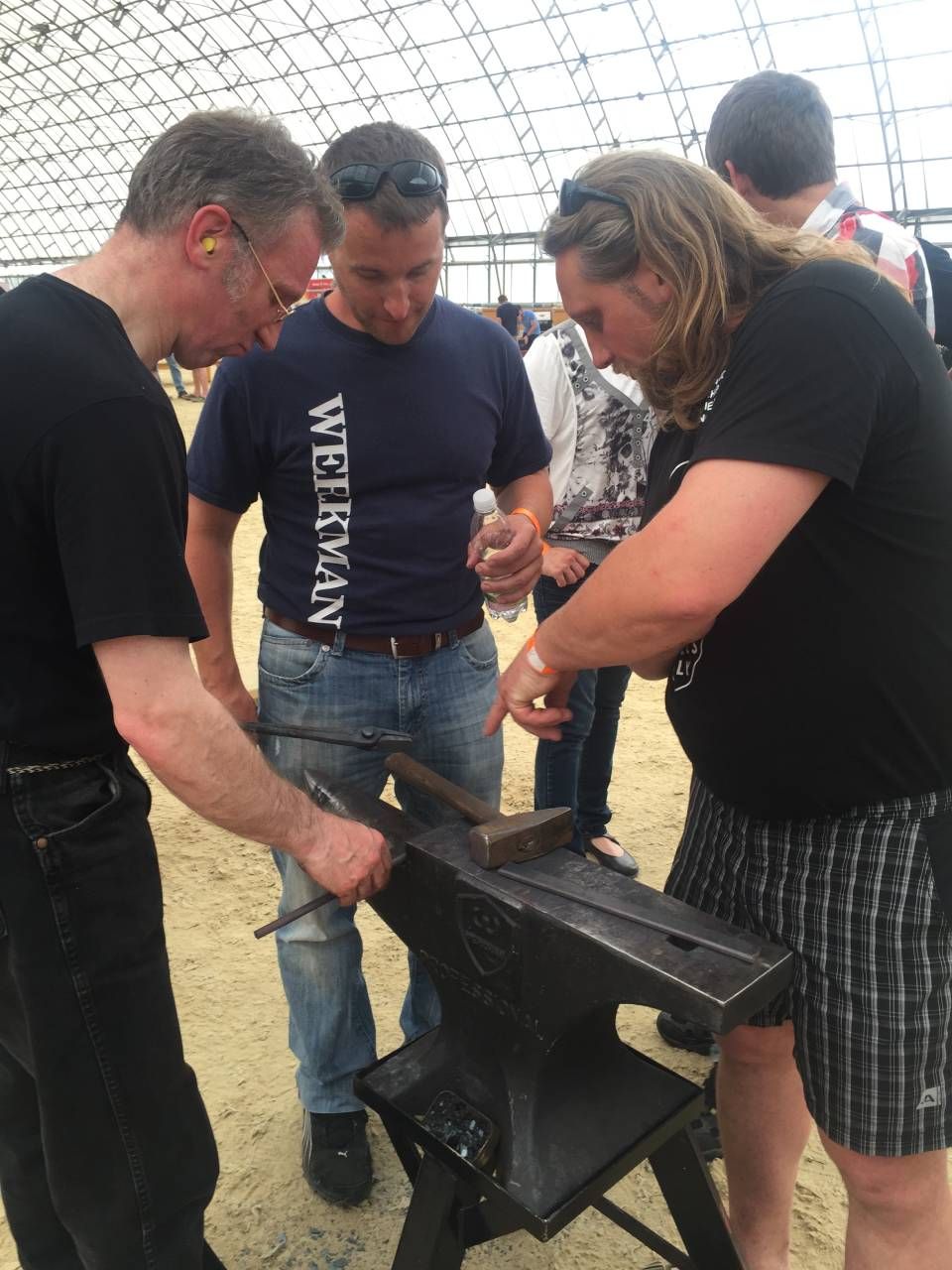 Drei Personen arbeiten an einem Amboss, eine hält einen Hammer. Die Szene spielt im Freien.