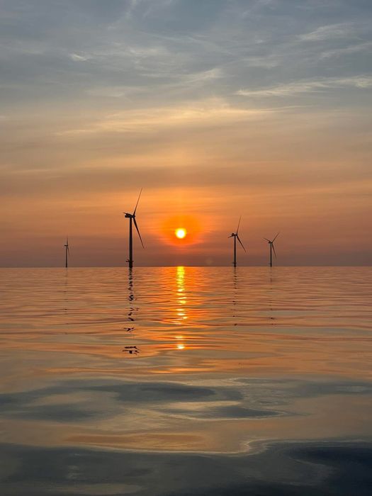 The sun is setting over the ocean with wind turbines in the foreground.