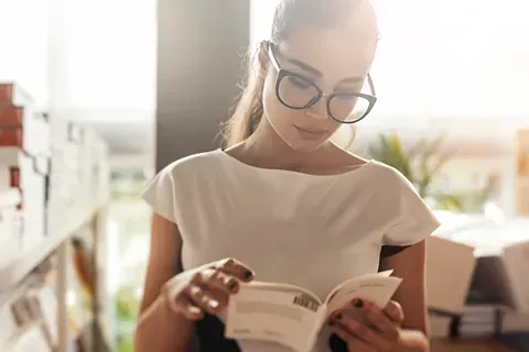 Una mujer que lleva gafas está leyendo un libro en una biblioteca.