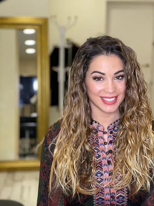 Una mujer con cabello largo y rizado sonríe frente a un espejo.