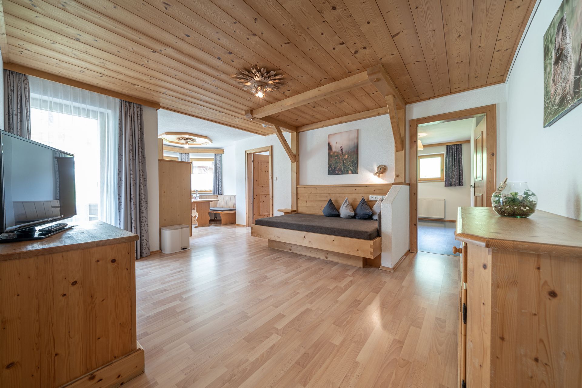 Ein geräumiges, holzgetäfeltes Hotelzimmer mit Fernseher, einer Couch, die auch als Bett dient, hellen Holzböden.