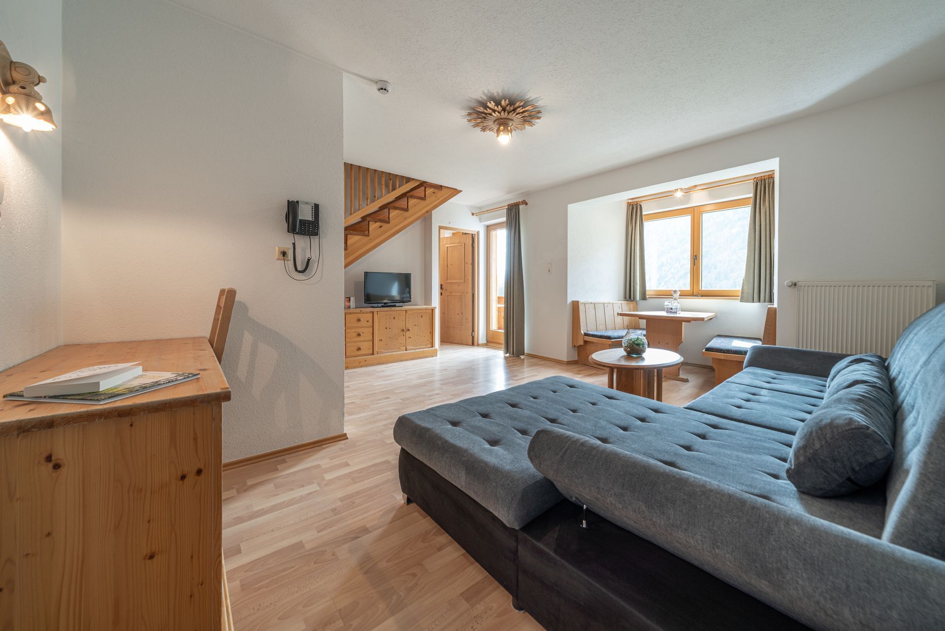 Ein möbliertes Hotelzimmer mit Schlafsofa, Holzschreibtisch, Fernseher, das durch ein Fenster mit Tageslicht durchflutet wird