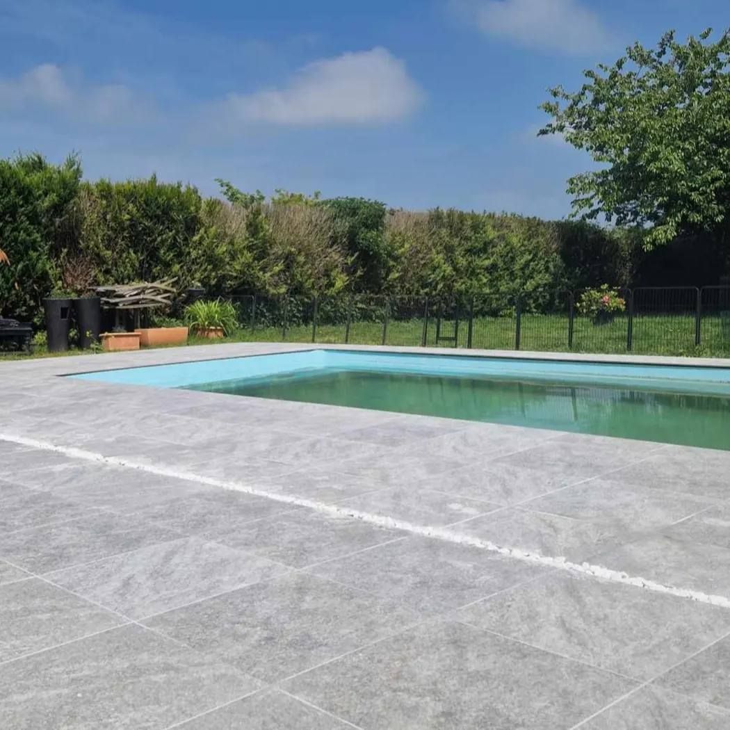 Piscine avec terrasse en pierre grise, eau verte et clôture bordée d'arbres sous un ciel bleu
