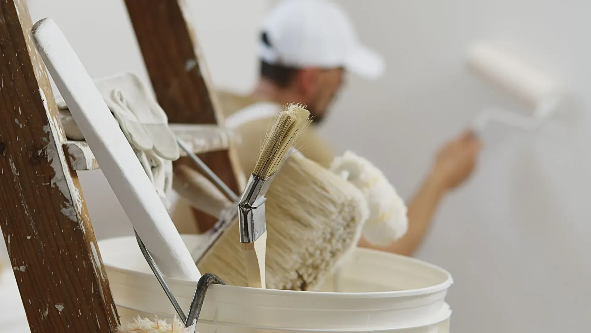 Peintre en chapeau blanc utilisant un rouleau sur un mur blanc, pot de peinture avec pinceaux, échelle.