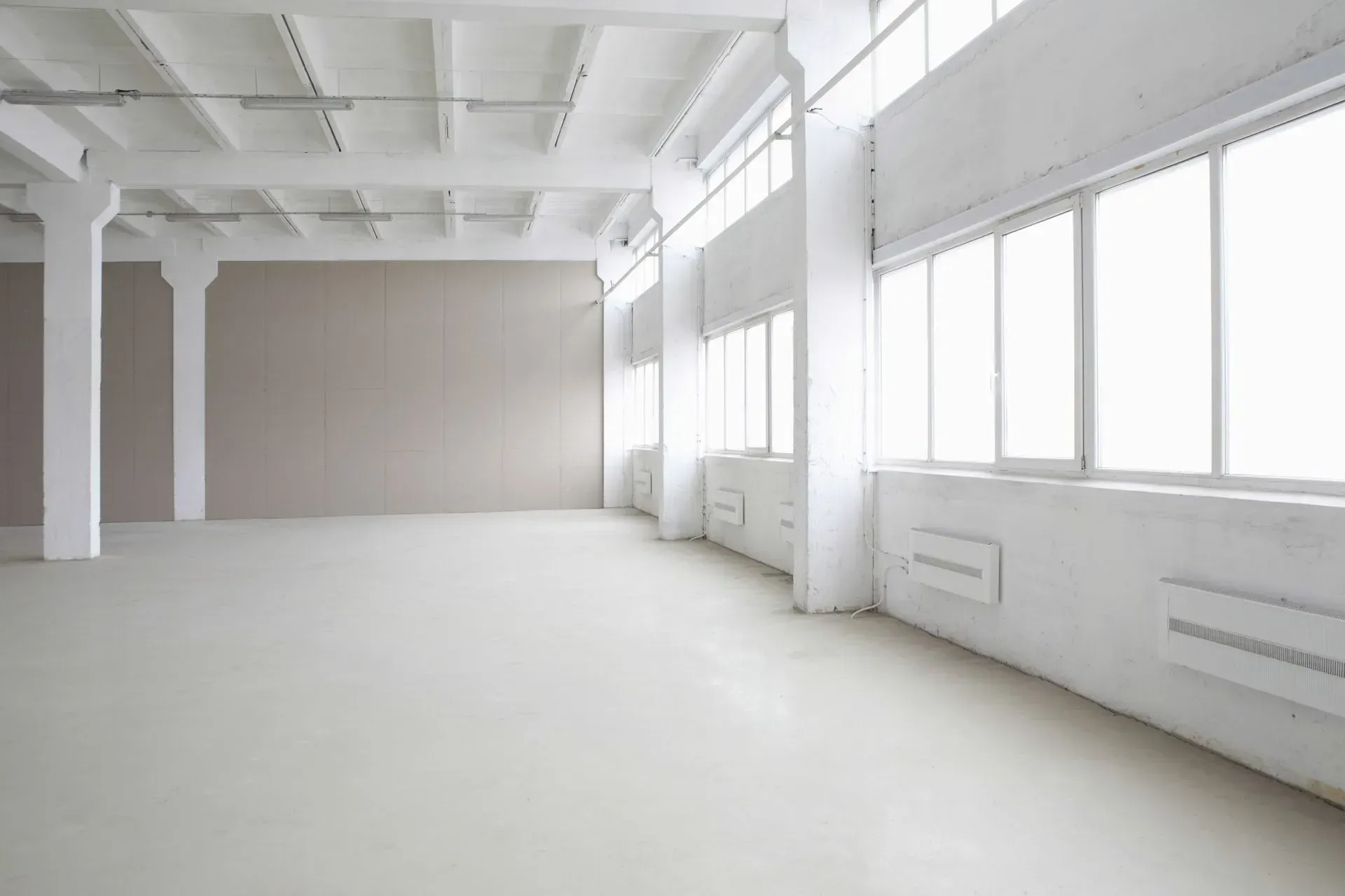Espace industriel vide et blanc lumineux avec de grandes fenêtres et des piliers en béton.