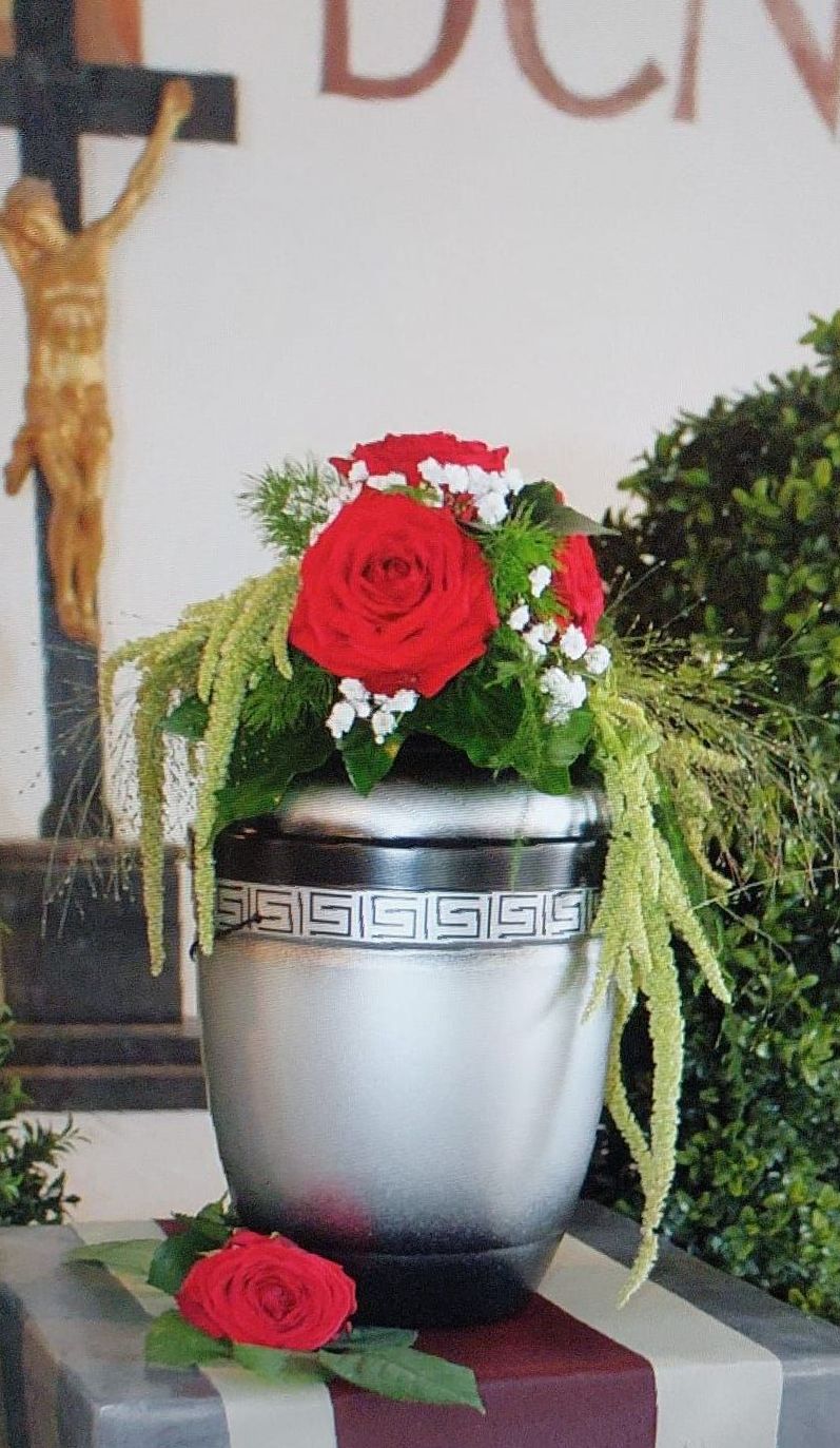 Eine silberne Vase mit roten Rosen steht auf einem Tisch vor einem Kreuz.