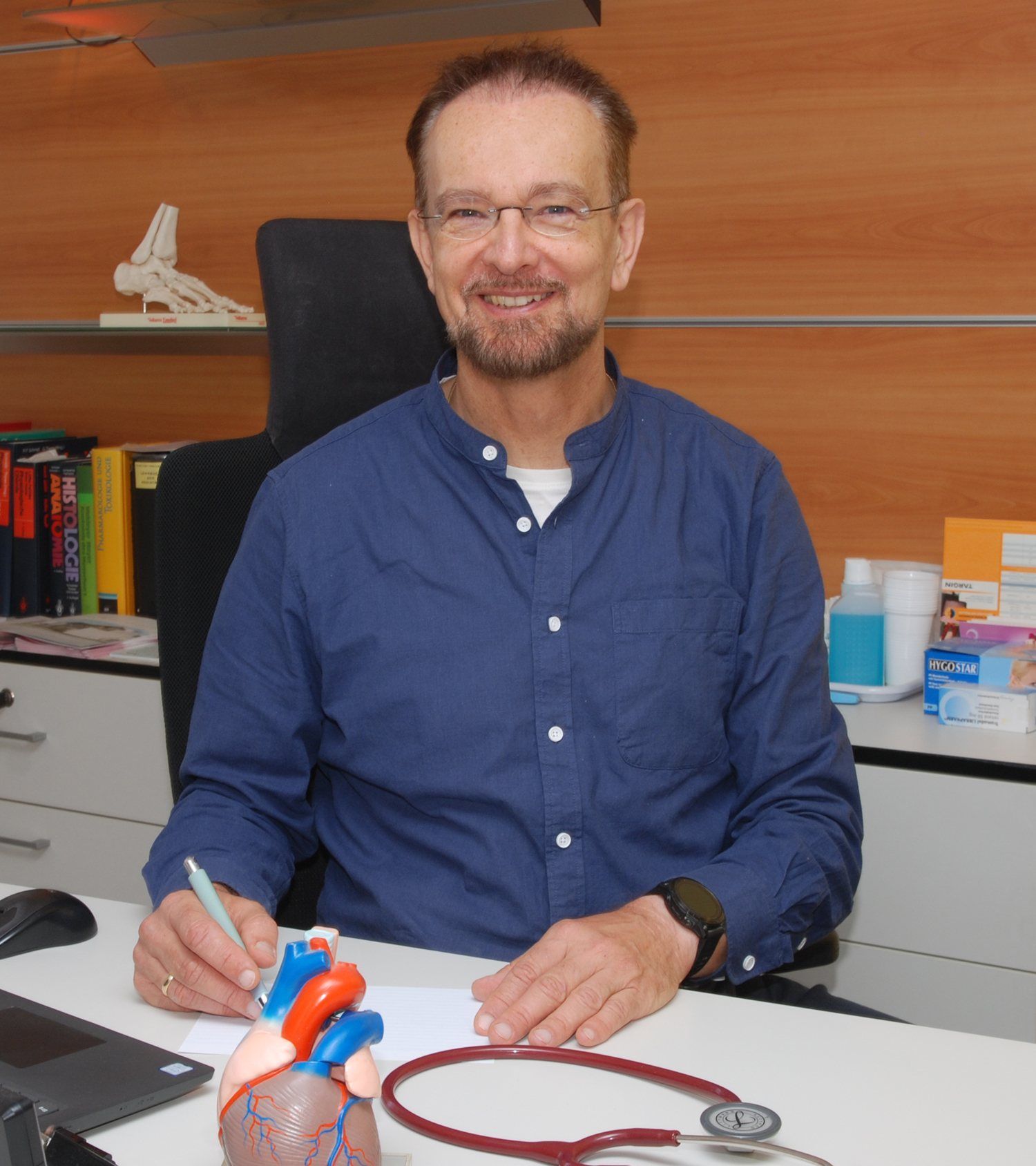 Mann im blauen Hemd lächelt an einem Schreibtisch mit einem Modellherz und einem Stethoskop.
