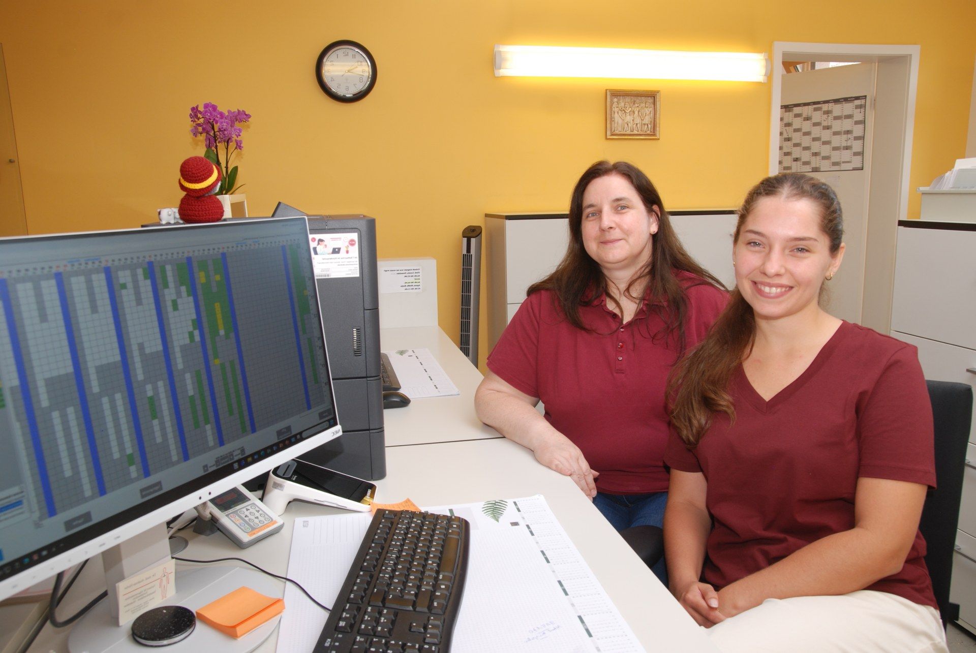 Zwei Frauen in bordeauxroten OP-Kitteln sitzen lächelnd neben einem großen Monitor in einem Arztzimmer.