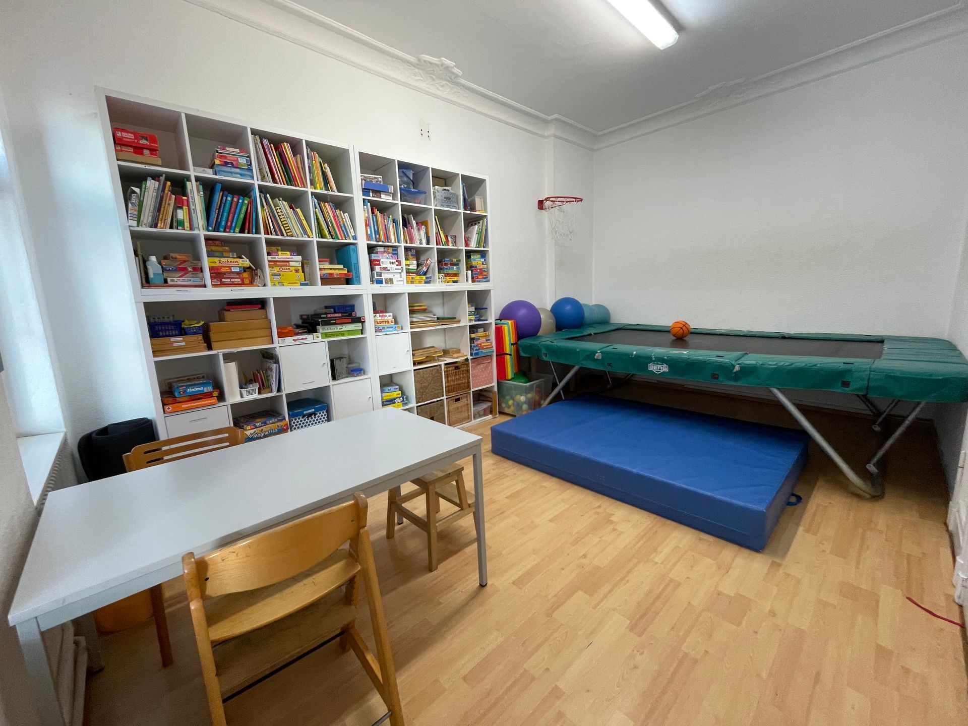 Kleines Spielzimmer mit Trampolin, Gymnastikbällen, Hockern, Schränken und einem Tisch auf Holzboden.