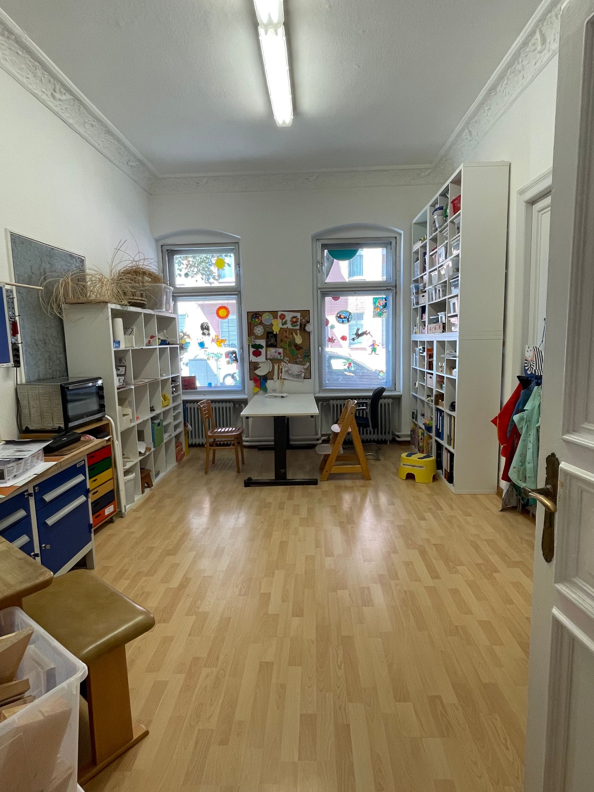 Zwei Personen sitzen an einem Tisch in einem hellen Klassenzimmer mit Holzschränken und bunten Aufbewahrungsboxen.