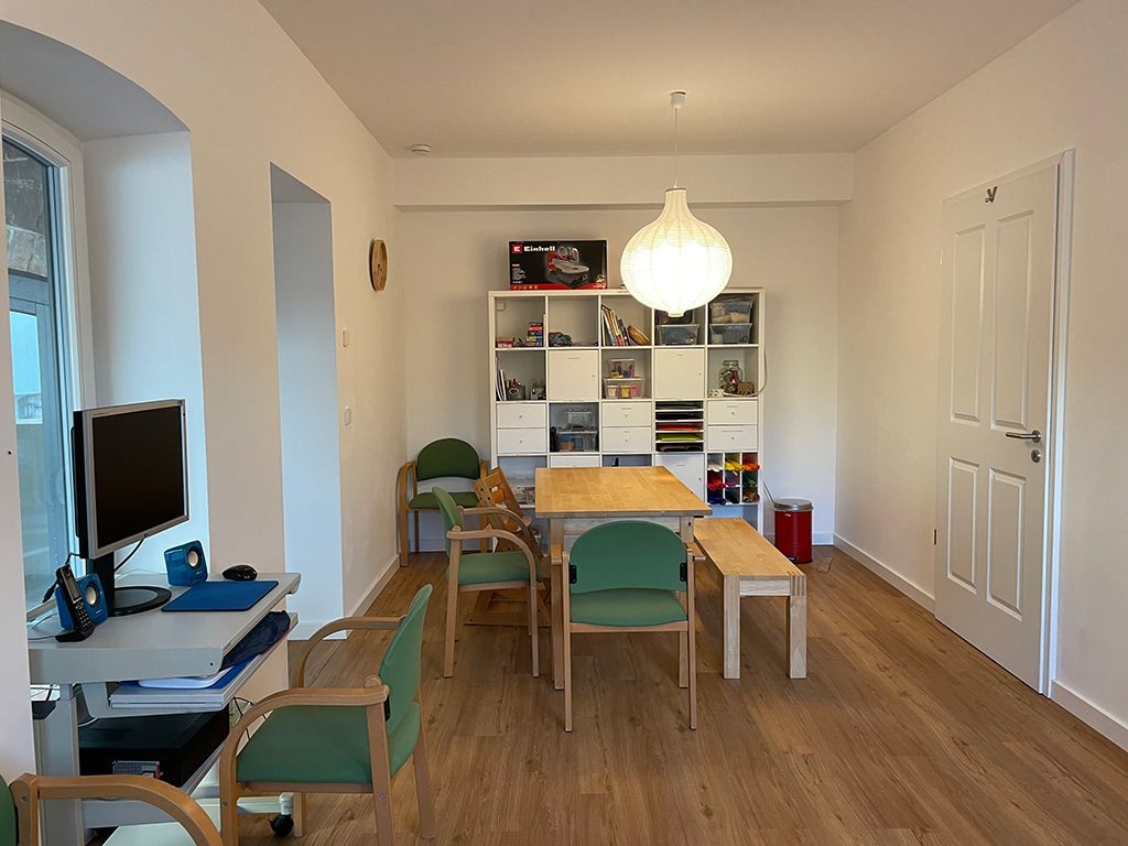 Helles Esszimmer mit Holztisch, grünen Stühlen, Bücherregal und Fernseher am Fenster.