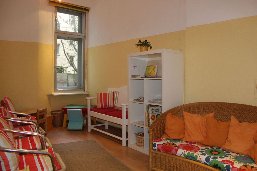 Gemütliches Wartezimmer mit gestreiften Stühlen, einem kleinen Tisch, einer Couch und weißen Regalen am Fenster.