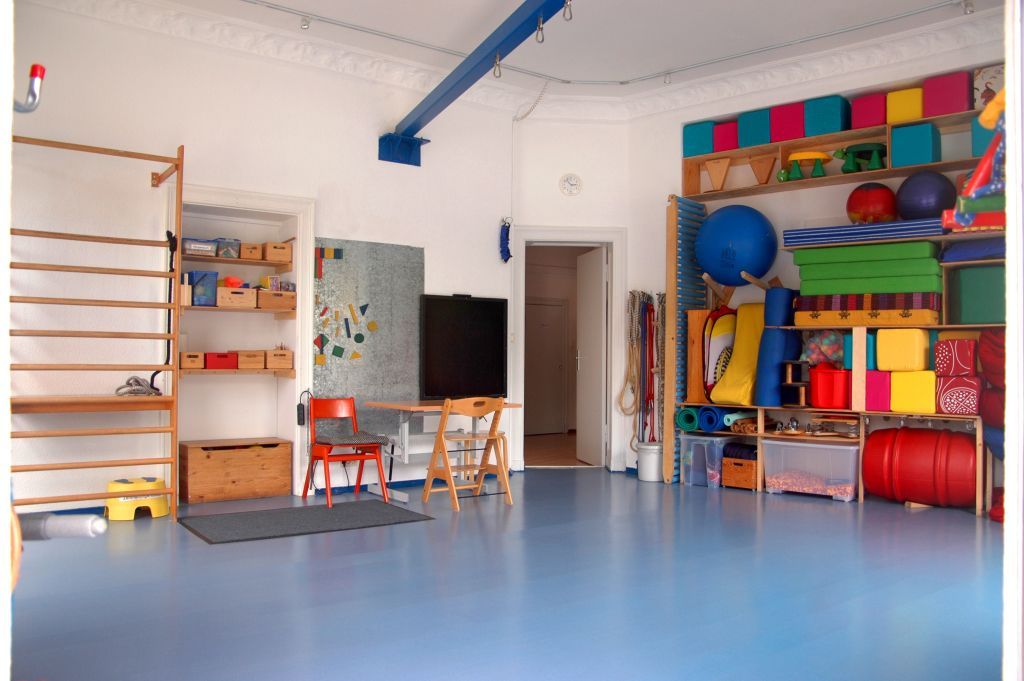Farbenfrohes Kinderspielzimmer mit Kletterstangen, Regalen, Schaumstoffmatten und kleinen Tischen auf blauem Boden.