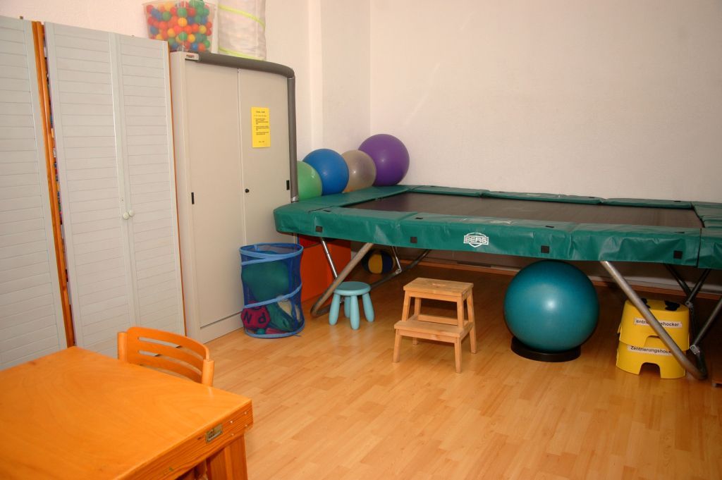 Kleines Spielzimmer mit Trampolin, Gymnastikbällen, Hockern, Schränken und einem Tisch auf Holzboden.